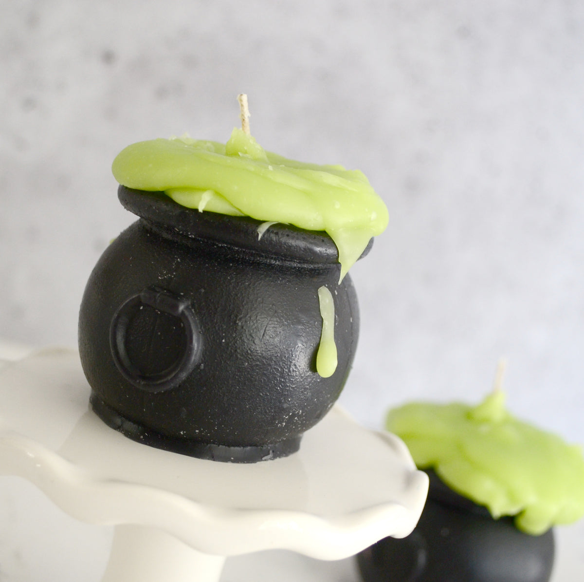 Black cauldron-shaped candle with green wax on a white stand against a gray background