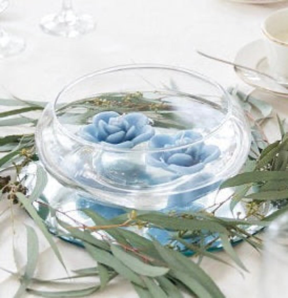 A bowl of floating rose shapes candles sits on a round mirror surrounded by eucalyptus leaves in the middle of a banquet table. 