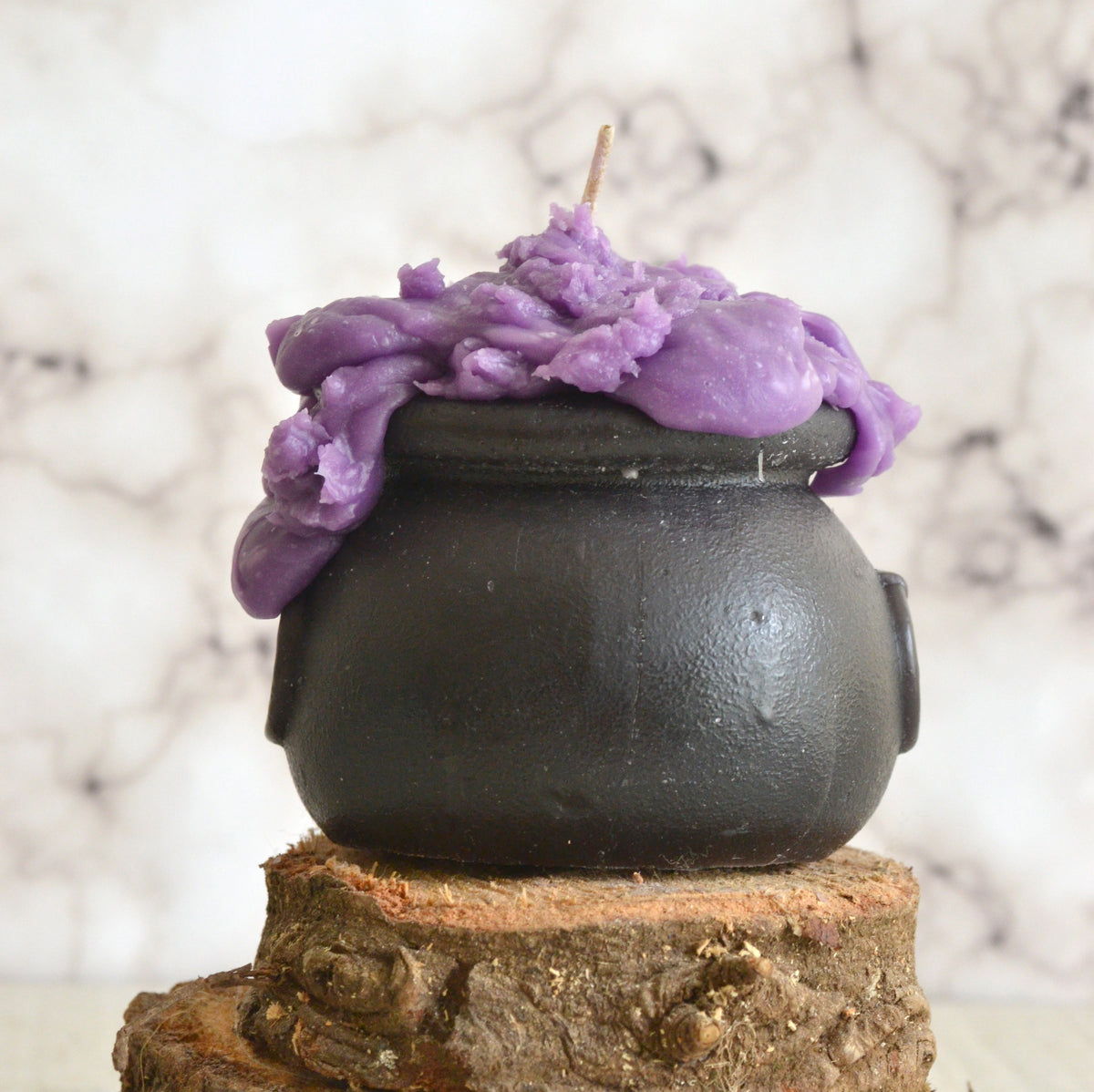 A purple soy wax candle with a black beeswax cauldron base, designed to resemble a witches' cauldron, placed on a wooden surface.